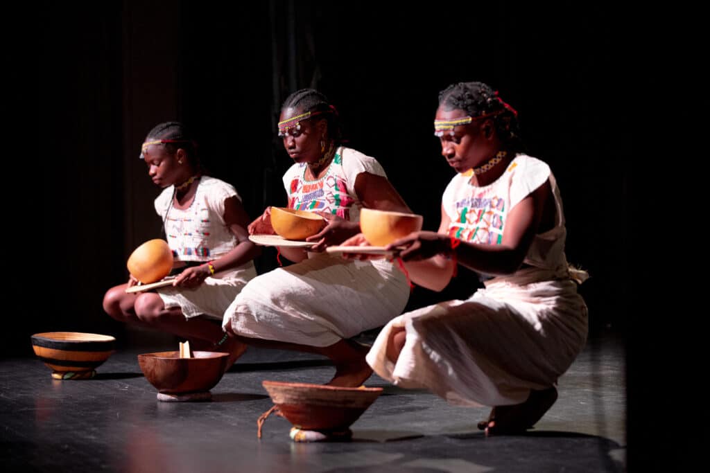 Performers kneel with calabashes under a single stage light in A Journey to Africa